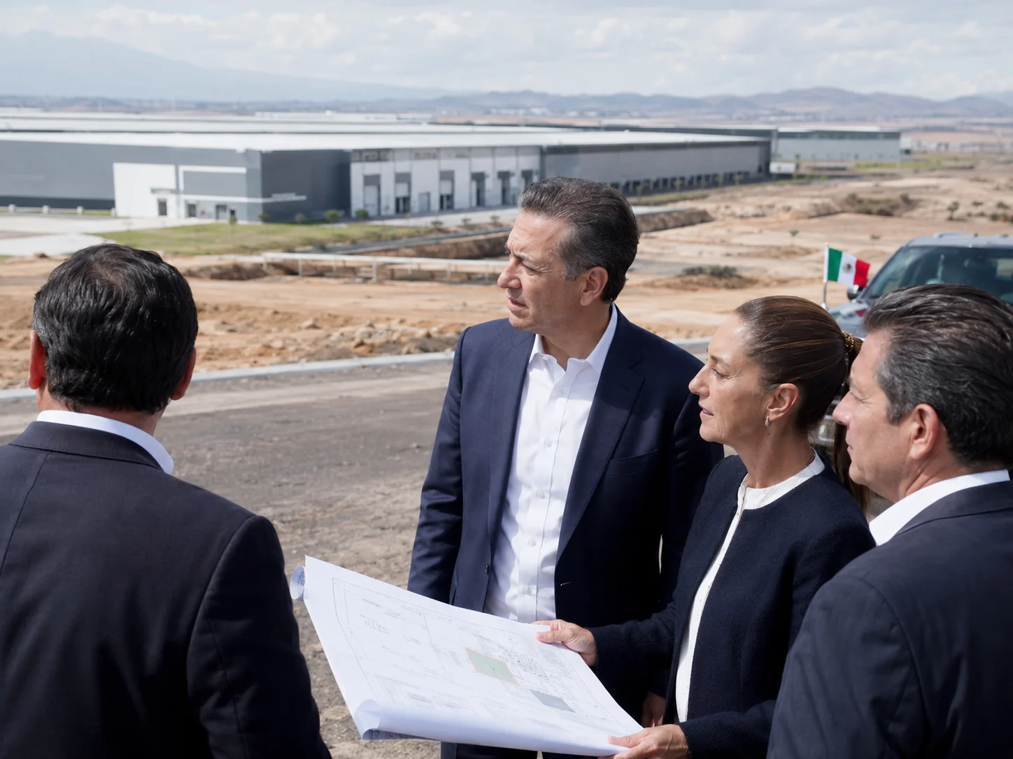 Funcionarios revisando planos frente a edificio industrial moderno con bandera mexicana.